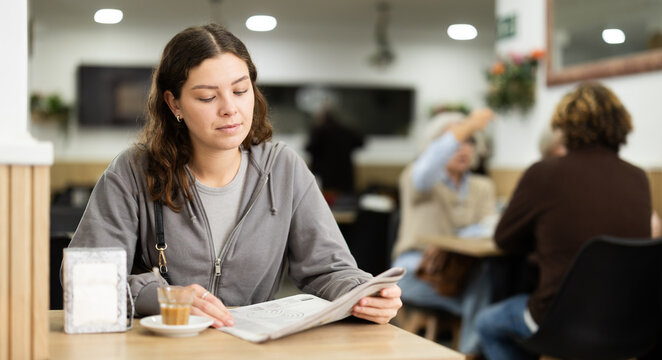 Attractive young girl sits alone at table in cozy cafe, drinks coffee and reads a newspaper
