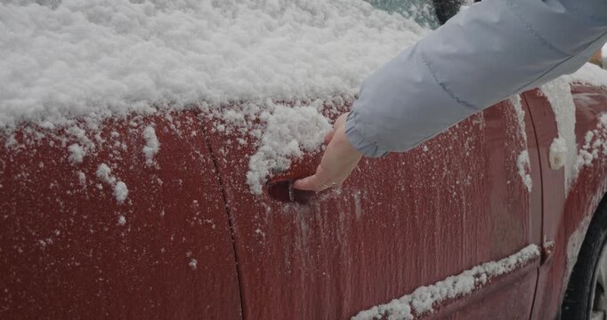 Close-up view of a person's hand unsuccessfully trying to open a red car door handle that is completely frozen and covered with a thick layer of ice and snow after a freezing rain storm.