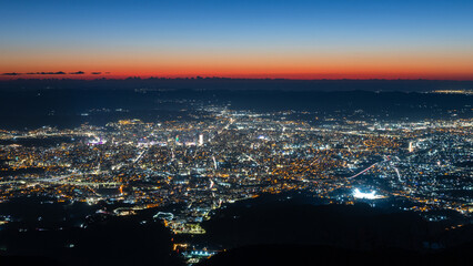 Naklejka premium Wide aerial view of a densely built capital illuminated after sunset with streets, neighborhoods and mountain silhouettes in sunset sky, Tirana, Albania
