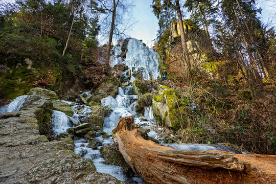 Wide angle view of ice covered and frozen Blauenthal waterfall with a fallen tree in the foreground in the Ore Mountains forest during winter season
