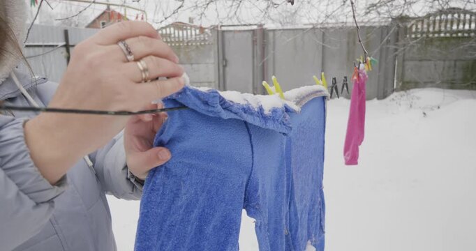Young woman in a winter coat taking down stiff, frozen laundry from a clothesline in a snowy backyard, demonstrating the effects of subzero temperatures on wet clothes during winter.