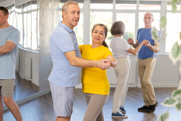 Positive senior woman and man practicing tango dance moves as a couple during a group celebration in a dance studio