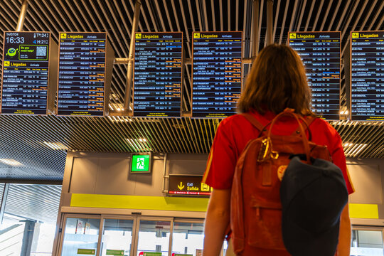 Young woman with backpack and cap on her back looks at the flight boards at the airport