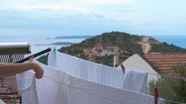 Person hanging clean white laundry on clothesline with sea view. Woman hangs freshly washed white sheets on a clothesline to dry outdoors on a balcony. The beautiful background shows a coastal village