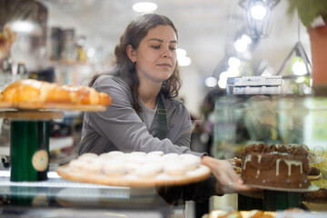 Positive young woman seller putting fresh pastries on plate from counter in small cafe bakery