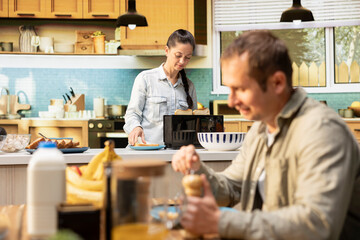 Obraz premium Wife preparing eggs at counter for her husband to eat breakfast together at table, serving scrambled eggs with bacon and pastry. Cozy scene captures bonding with joy for morning routine.