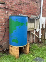 Blue rainwater barrel connected to house gutter, standing on bricks beside a brick wall, simple outdoor rainwater harvesting system for garden use.