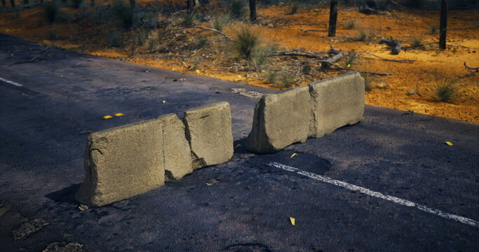 concrete barriers blocking rural road, cracked asphalt, golden dry grass, scattered leaves, roadside trees in warm dusk light makeshift blockade suggests human