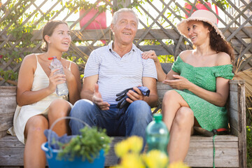 Obraz premium European woman in straw hat sitting on wooden bench, chatting sweetly with sister and elderly father. Rustic setting, young women and gray-haired man on background of wooden trellis discussing news