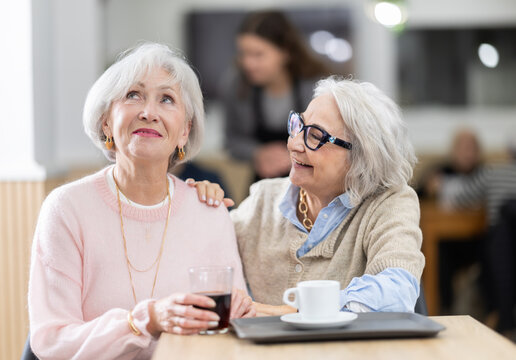 Two senior women colleagues meeting and drinking coffee at table in small cafeteria