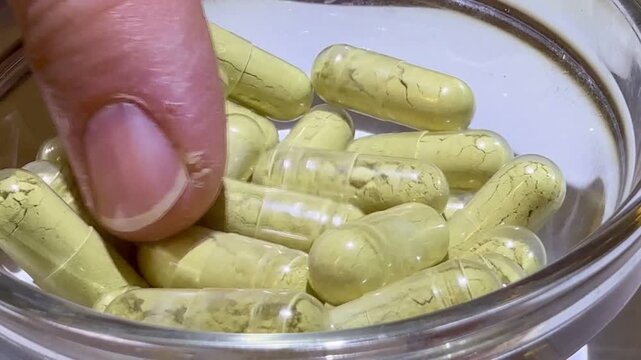 Close-up of woman's fingers counting capsules in glass jar, soft clinical lighting, beige herbal capsules with visible granules, careful fingertip movement, dosing routine and supplement