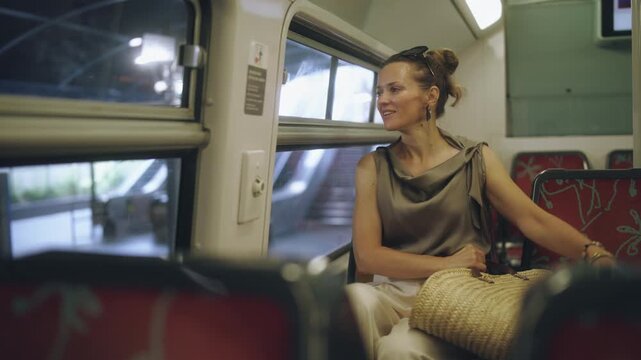 A smiling woman enjoys the view from a train window while traveling through Paris, holding a straw bag and dressed in a stylish summer outfit.