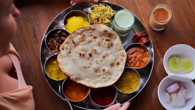 Top view of Indian thali meal with multiple traditional dishes including rice, curries, pickles, papad, chutney, and sweet, served on a steel plate with two hands interacting, showcasing Indian culina