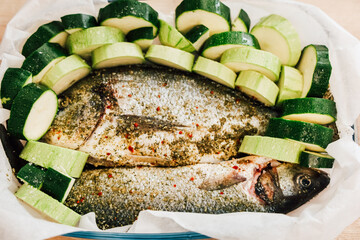 Fresh dorado fish and sliced zucchini prepared for baking in a dish with herbs and spices