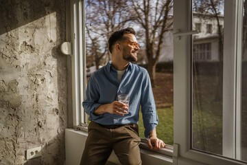 Happy man in casual wear drinking water at window