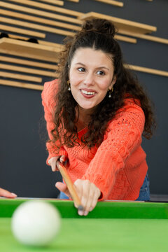 Friends at green-felt pool table, female in orange sweater leaning, aiming cue stick at white ball