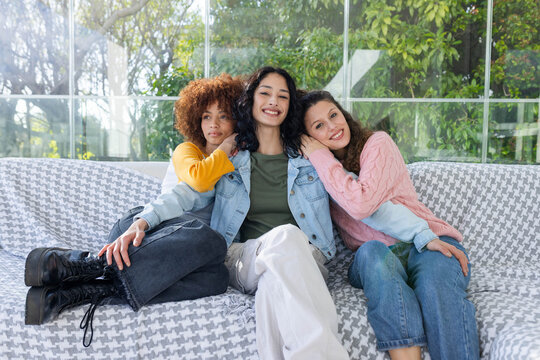 Diverse female friends sitting on houndstooth throw couch by glass panels, wearing denim jacket