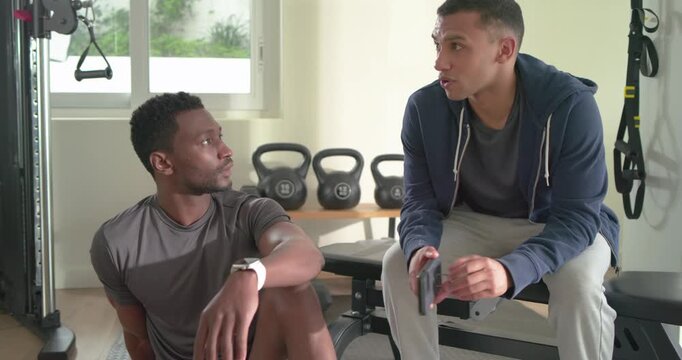 Diverse male partners tapping smartphone sharing fitness tips sipping water by bench in home gym