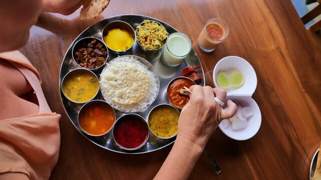 Top view of Indian thali meal with multiple traditional dishes including rice, curries, pickles, papad, chutney, and sweet, served on a steel plate with two hands interacting, showcasing Indian culina