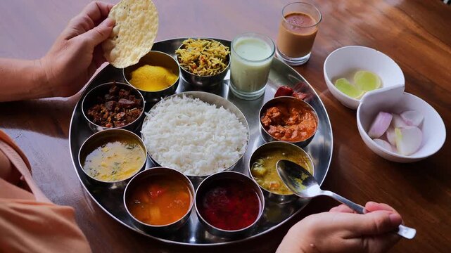 Top view of Indian thali meal with multiple traditional dishes including rice, curries, pickles, papad, chutney, and sweet, served on a steel plate with two hands interacting, showcasing Indian culina