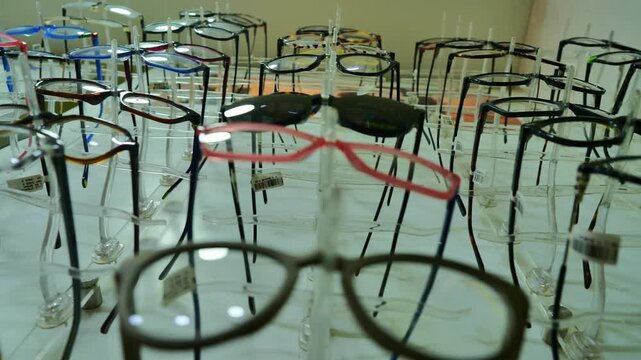Close up view of assorted eyeglass frames displayed on clear stands inside optical store, showing variety of colors and shapes, organized retail presentation, vision care shopping environment, and mod