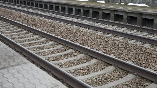 Railway tracks extend into distance.  Gravel fills spaces between rails.  Concrete barriers border the scene.  Snow lingers atop wall.  Motion shows progression along route