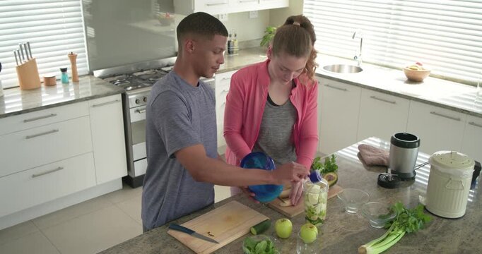 Couple preparing smoothie man pouring blue pitcher into jar while woman slicing banana in kitchen
