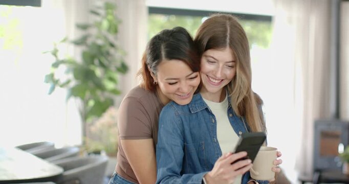 Diverse women standing close, second woman coming behind, leaning to view phone and mug, laughing