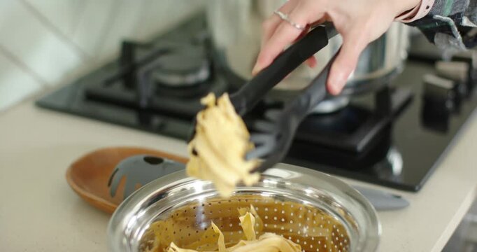 Hand with black tongs is lowering pasta into pot on stove with water boiling to cook