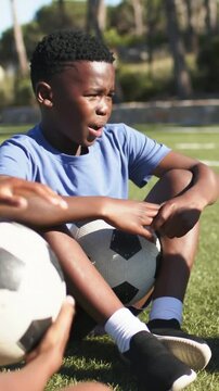 Vertical video: Watching pal bringing ball, blue youth sitting on field with b or w ball hearing coa