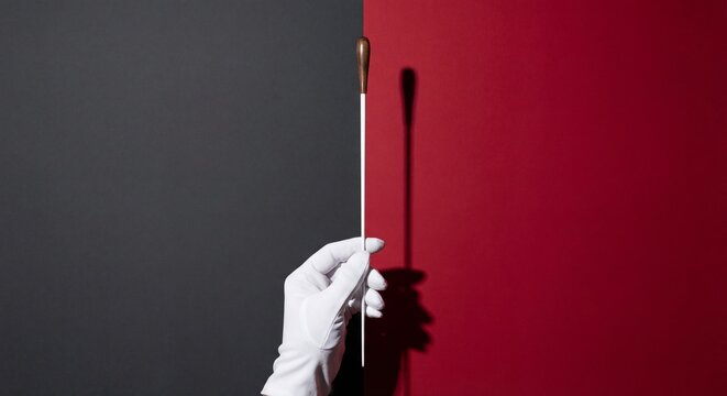 Hand in a white glove holding a conductor's baton on a split black and red background, representing leadership, precision, and musical excellence