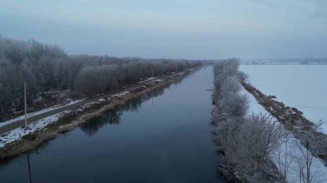 4K drone tracks the warm canal along the Konin area, with frozen fish ponds and snowy levees nearby. The shot emphasizes the thermal water corridor cutting through a deep-winter landscape.