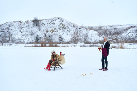 Elegant woman in fur coat and red boots sits in snowy field with a butler bringing her drinks, a surreal winter scene blending luxury and whimsy against a backdrop of natural beauty and subtle charm.