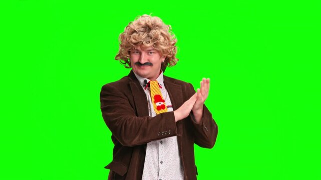 A man wearing a blond curly wig, a brown jacket, a bright tie and a mustache claps his hands with a humorous expression while standing in front of a green screen.