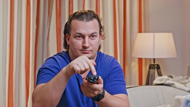 A man in a blue shirt sits on a sofa holding a remote control in his hands, pointing forward, frantically switching channels while sitting in a cozy room
