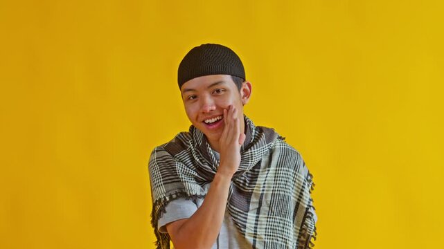 Young Asian Muslim man whispering secret with hand near mouth and wearing black skullcap and keffiyeh scarf isolatedon yellow background