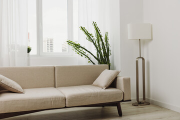 Minimalist living room with beige sofa and matching cushions under large bright windows. Tall green...