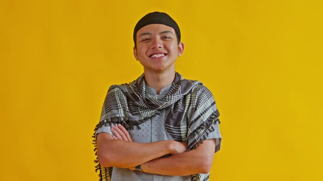 Smiling young Muslim man wearing peci and checkered scarf crossing arms confidently on yellow backdrop