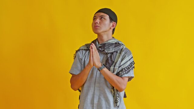 Peaceful Muslim man wearing peci and scarf praying and seeking guidance against vibrant yellow backdrop
