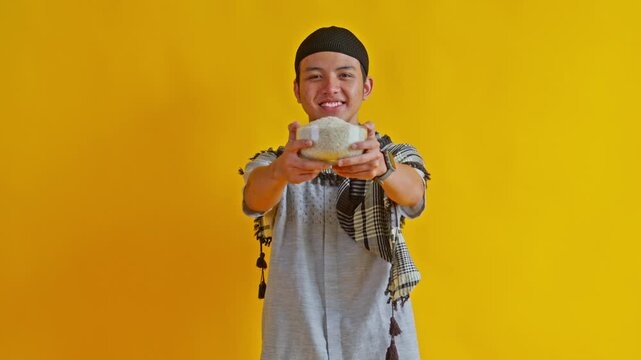 Smiling young Asian Muslim man holding and offering bowl of rice for Zakat charity against yellow background
