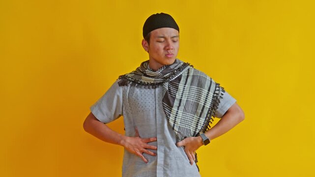 Young Asian Muslim man checking wristwatch and holding stomach while waiting iftar impatiently against yellow background
