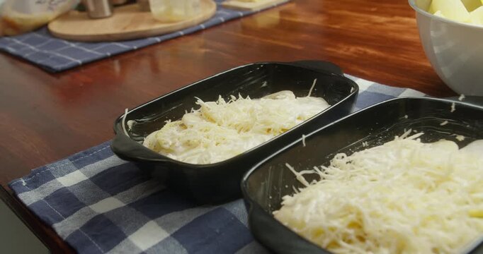 Chef pours creamy white b chamel sauce over layered sliced potatoes and cheese in cast iron baking dishes. Close-up of cooking process, homemade comfort food, classic French sauce,