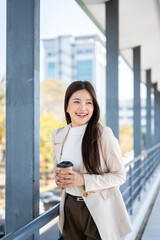 Fototapeta premium Pretty asian woman office worker holding coffee with both hands as standing or walking on footbridge