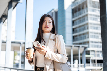 Obraz premium Asian woman office worker holding phone and coffee as standing walking on footbridge with cityscape.