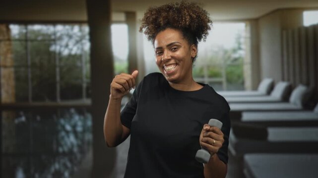 Woman young african american smiling and holding dumbbell in spa building lounge with poolside beds and large windows; joyful fitness.