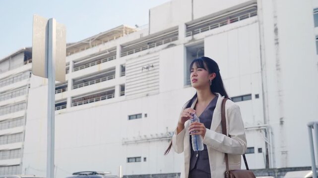 Young businesswoman walking outdoor street, shielding eyes from sun, holding water bottle, looking up at modern office buildings, enduring hot weather during city travel, professional career path.