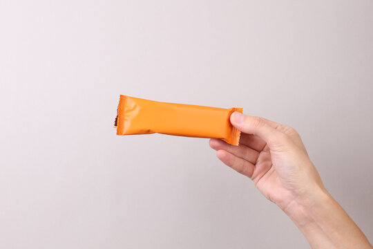 Hand holding Chocolate bar pack mockup on gray background. Blank orange candy wrapper