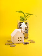 Mini house model with coin jar and plant growing, symbolizing real estate investment, personal finance, asset growth, long term planning, and sustainable financial future on vibrant yellow background.