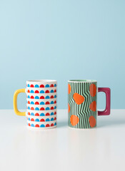 Two Colorful Ceramic Mugs with Abstract Patterns on White Surface Against Blue Background