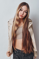 Fototapeta premium Young woman with long straight hair wearing beige cropped top and jacket posing in studio with neutral expression on white background. Casual style and fashion portrait.